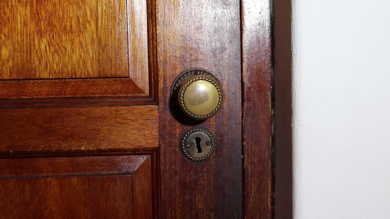 Man with a black glove on slowly opens a brass door knob on a wooden door