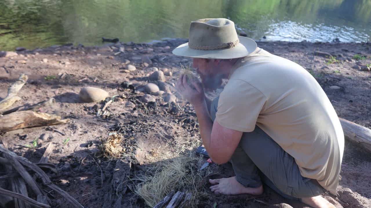 아쿠브라 모자를 쓴 부시맨이 호주 덤불에 있는 풀에 엠바를 날려 불을 피웁니다.