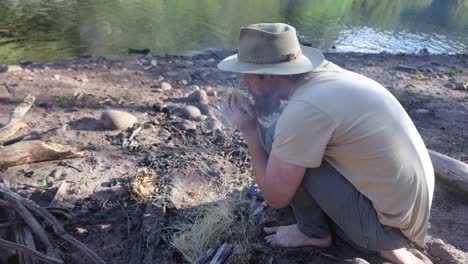 a bushman in an akubra hat creates fire by blowing an embar in grass in the australian bush