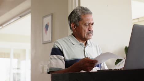 Biracial-senior-man-doing-paperwork-and-using-laptop-at-home,-slow-motion