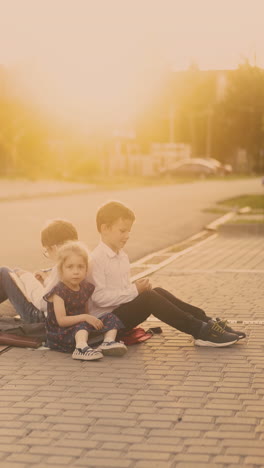 nervous schoolboy in white sits back to back with mate by sister throws textbook on asphalt road lit by back sunlight