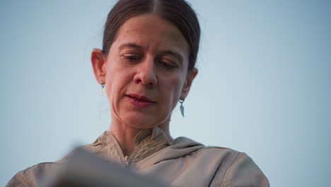 close up of middle age woman concentrating on writing in journal with calm focused expression outdoors against clear sky wearing beige jacket and leaf earrings