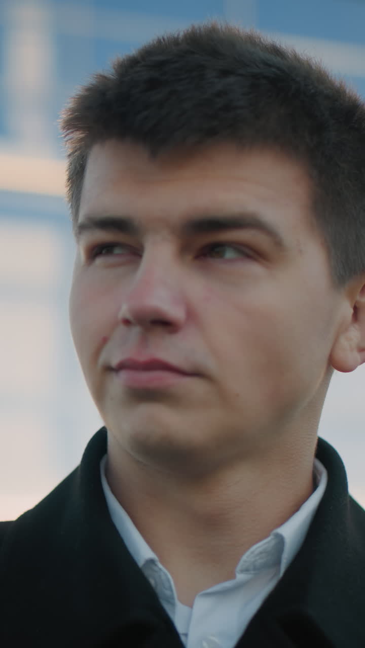 Business man in black clothing gazes into distant with a warm smile, light illuminating his left shoulder, background features modern urban building