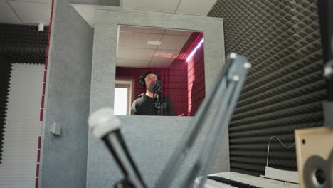 a man dressed in black stands in front of a studio microphone, wearing headphones as he sings passionately in a red soundproofed room. a recording session in a professional studio environment