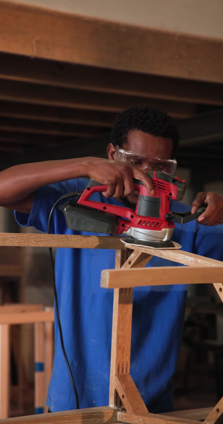 Man in blue shirt placing red sander, sanding frame for finishing in woodshop, coworker inspecting