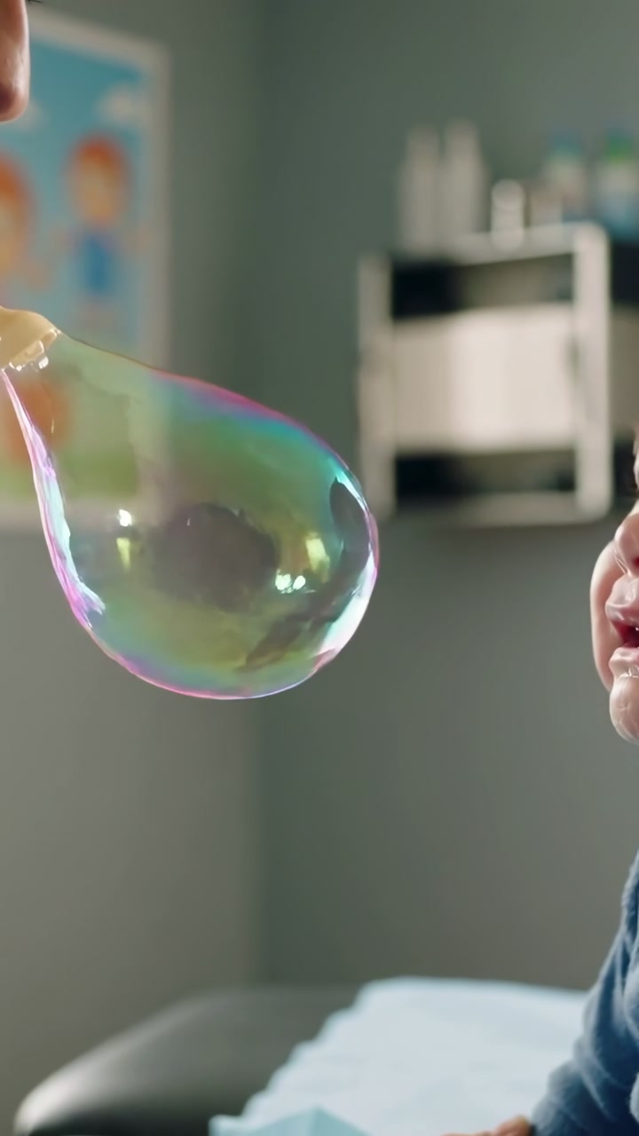Vertical video: Bringing wand, adult at clinic blowing bubble soothing toddler blue top, copy space