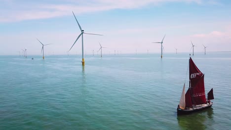 A-flat-bottomed-sailing-barge-sailboat-moves-up-the-Thames-River-Estuary-in-England-amidst-numerous-wind-turbine-windmills-4