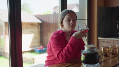 young adult in red blouse gently sips wine from glass while sitting indoors at wooden table near kitchen window, natural daylight streaming in, creating warm and relaxed atmosphere
