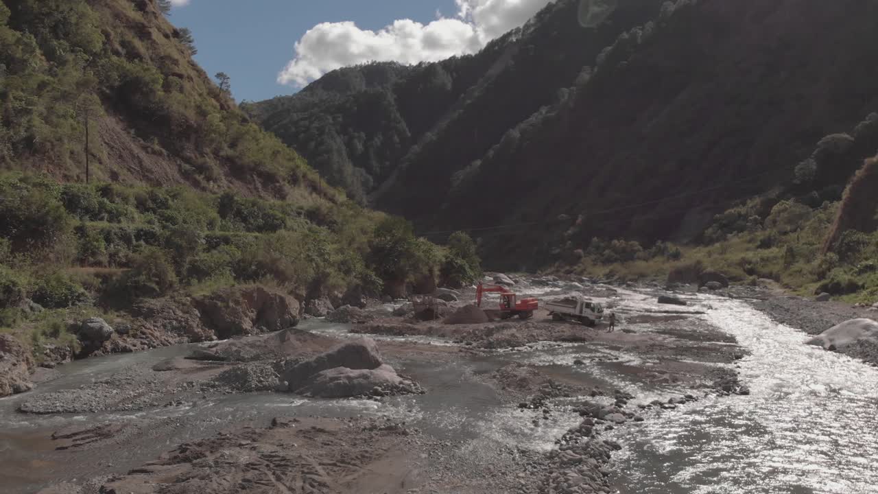 excavator digging rocks out of river bed in mountainous region in Benguet Kabayan Philippines wide angle aerial approaching raw cement material mining flowing water reflecting light bright sunny day