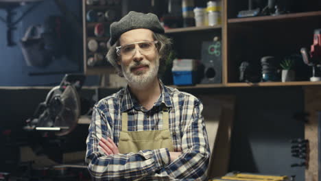 portrait of old caucasian handicraftsman in hat, apron and goggles looking at camera in carpentry workshop