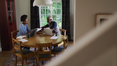 african american couple sitting at dining table using laptops paying bills