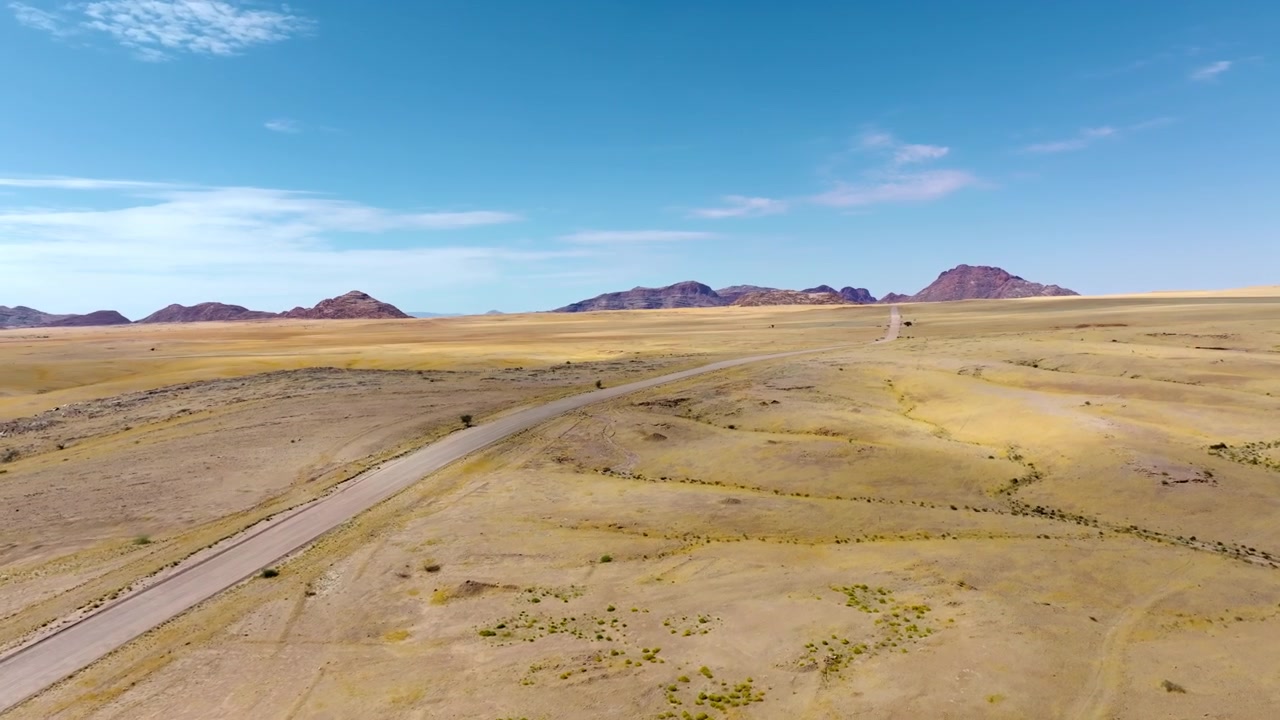 Drohnenansicht einer Schotterstraße, die sich über goldene Ebenen auf einen dunklen Granitberg in Namibia erstreckt. Goldene Graslandschaften ziehen sich unter einem strahlend blauen Wüstenhimmel auf markante rote Granitberge zu.