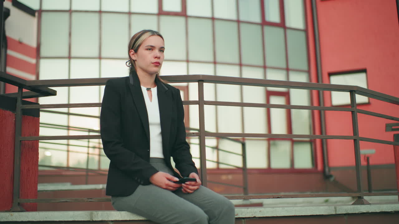 Pensive lady in black blazer and gray pants seated on fence looking at phone, waiting in urban environment with modern red and glass building