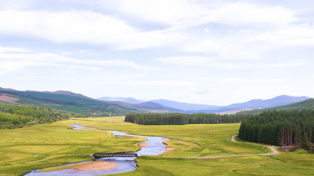 Drone glides above lush Scottish valley, river, bridge, meadows, forests, under soft daylight