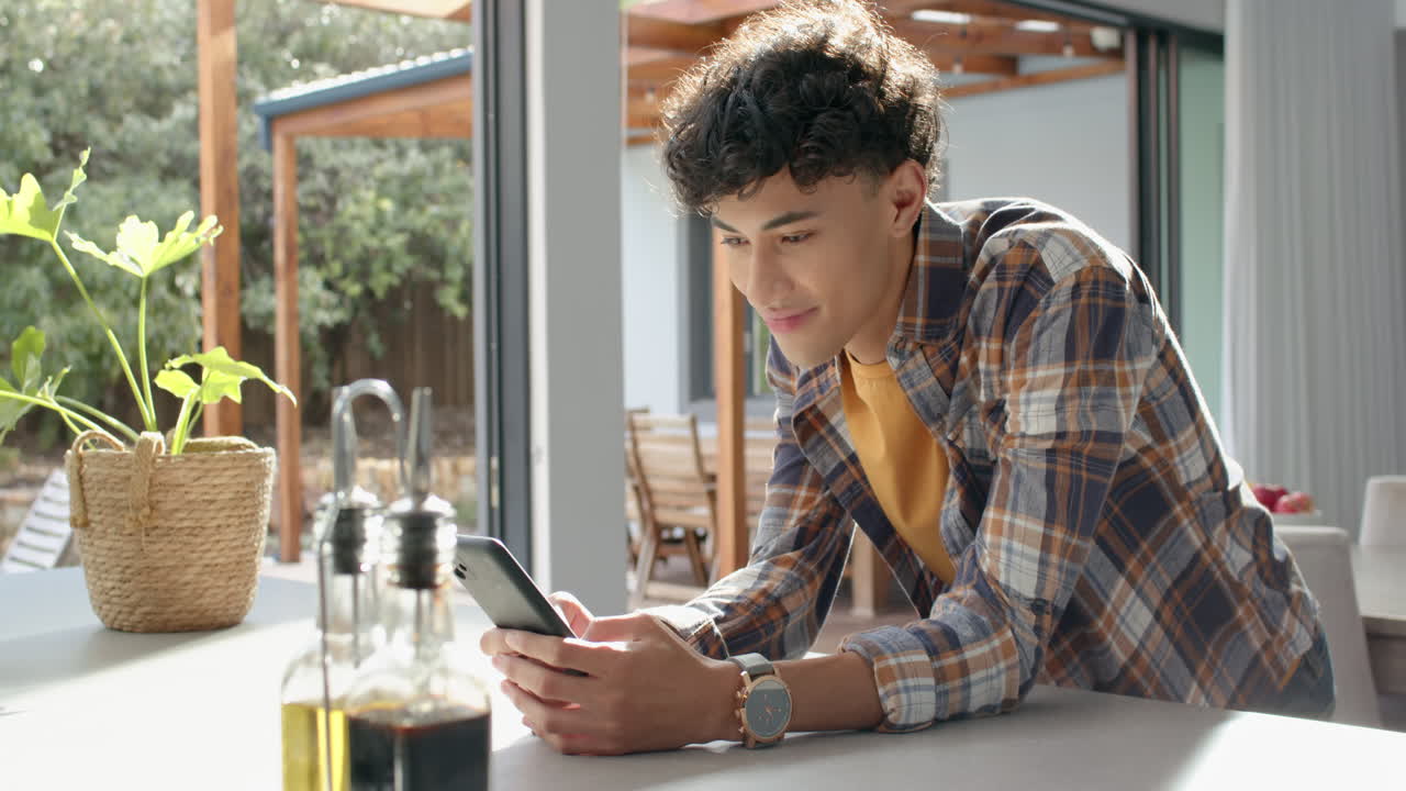 Smiling young man using smartphone while leaning on kitchen counter at home