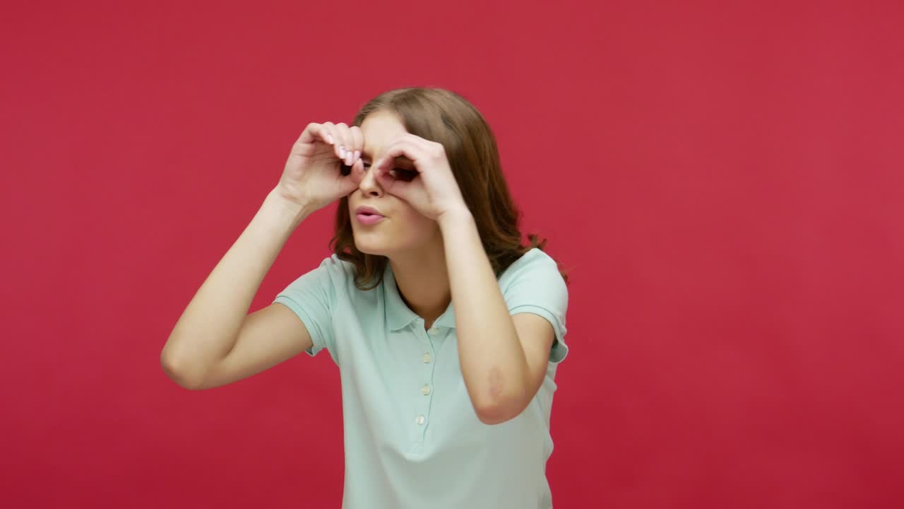 호기심 많은 젊은 여성이 손으로 안경을 만들고, 안경으로 손가락을 통해 몸짓을 봅니다.