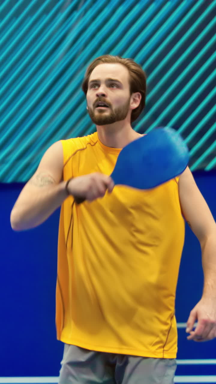 Man in yellow shirt playing pickleball on a blue, inside court. Vertical