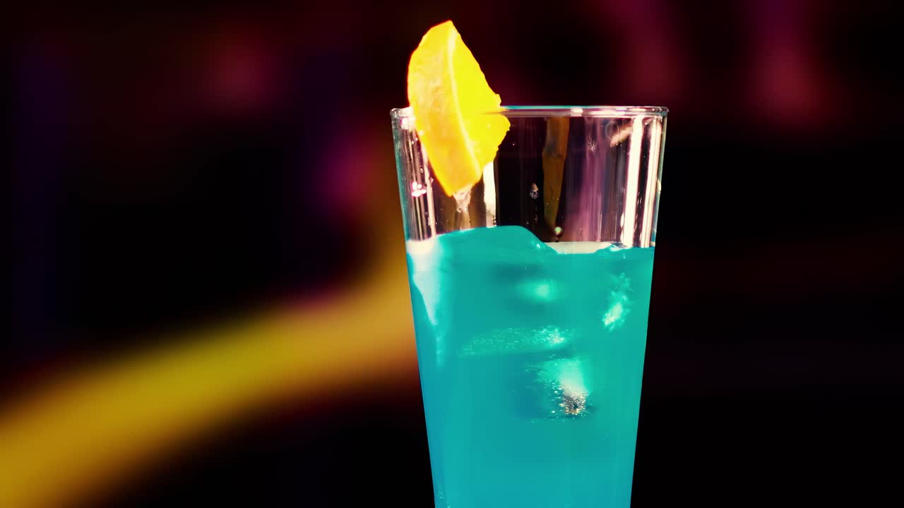 A blue drink with a lemon slice garnish against a dark, blurred background.