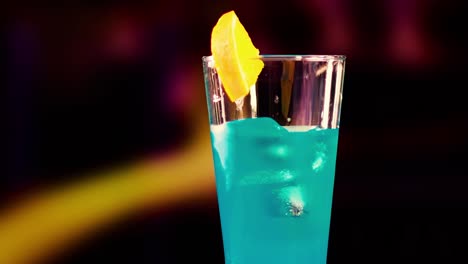a blue drink with a lemon slice garnish against a dark, blurred background.