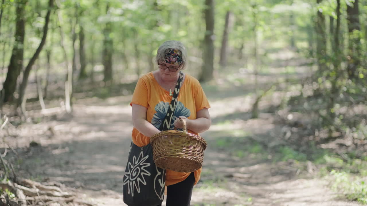 Inquadratura media di una donna anziana corpulenta con capelli grigi e maglietta arancione che guarda dentro il cesto di legno nel mezzo di una foresta verde durante una giornata di sole, al rallentatore e con sfondo sfocato.