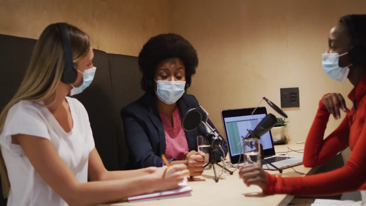 Three diverse female radio hosts wearing face masks talking on microphone for radio podcast
