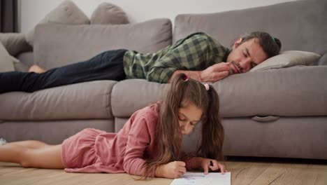 A-little-brunette-girl-in-a-pink-dress-draws-on-a-white-sheet-of-paper-with-a-pen-while-her-single-father,-a-brunette-man-in-a-green-checkered-shirt,-sleeps-on-a-gray-sofa