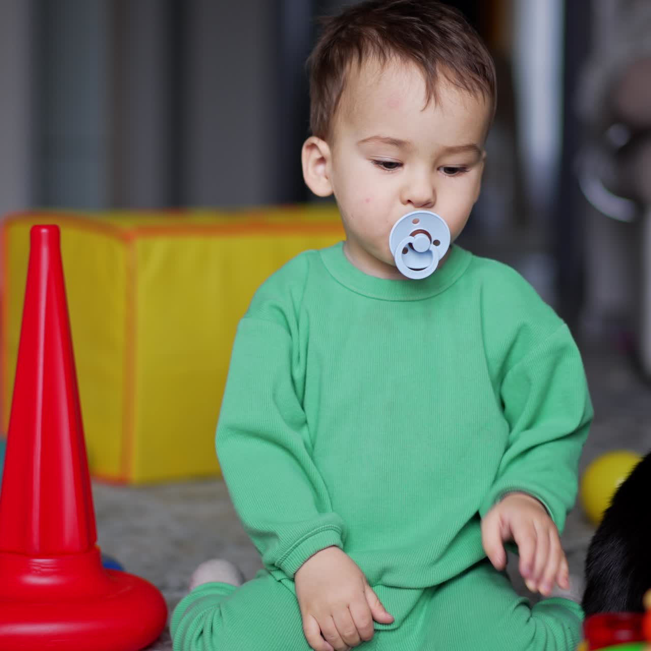 Calm lovely baby sitting on the carpet with toys around. Kid takes a pacifier out of mouth and takes back again. Black cat near the child
