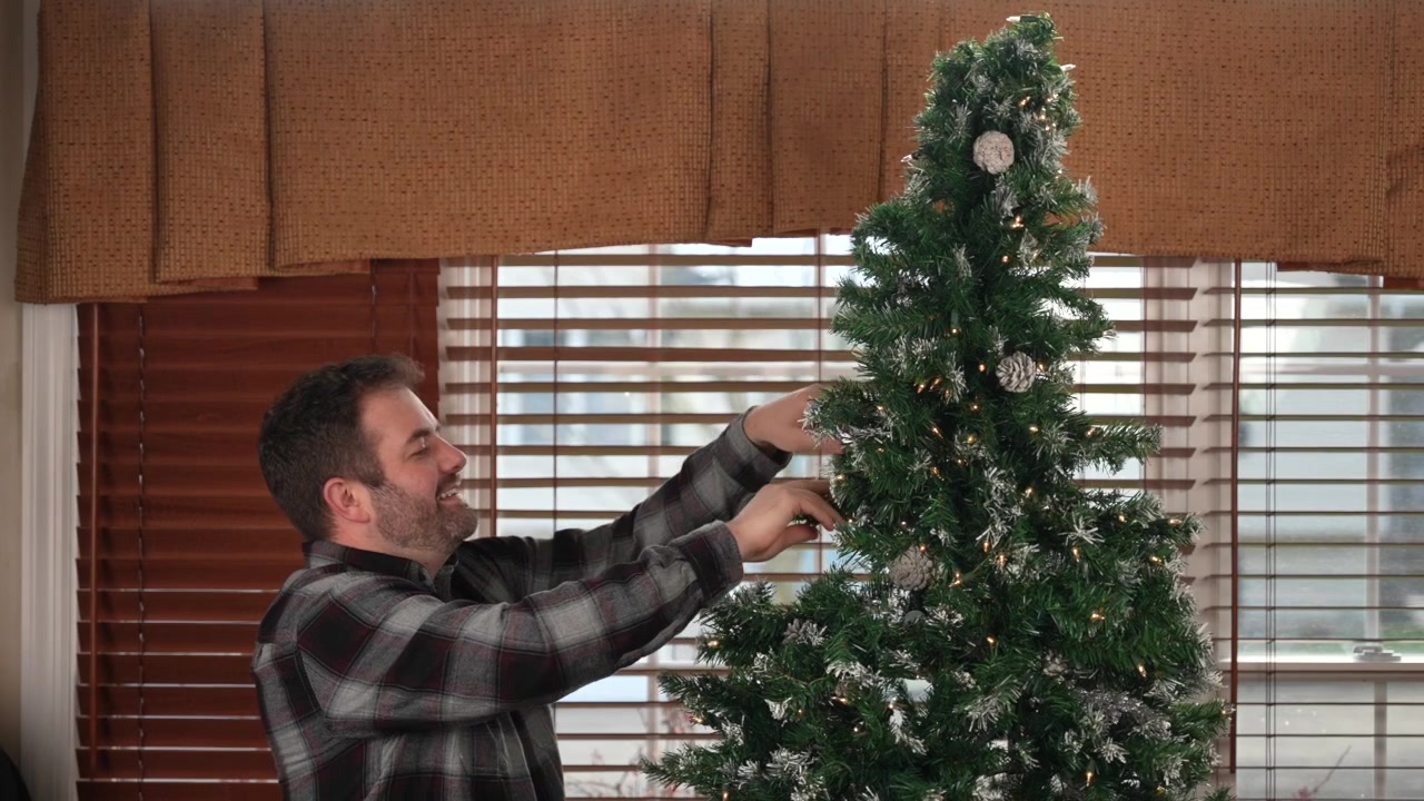 Um homem adulto coloca a peça final em uma árvore de Natal artificial, completando-a. A árvore se ilumina enquanto ele sorri, desfrutando do espírito festivo do feriado em 4K
