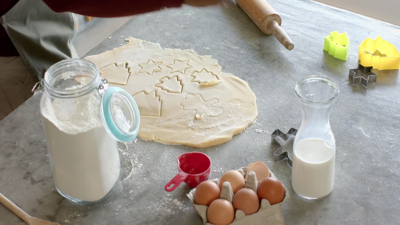 Baker's hands reaching for cutter, stamping star, tree shapes into dough on countertop for baking
