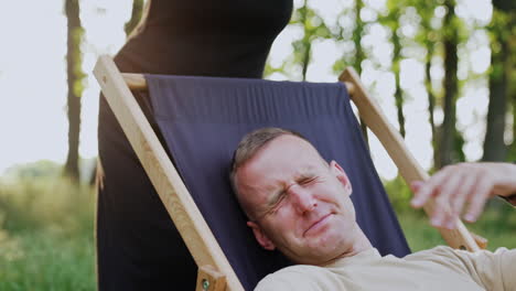 man slept in chair having face covered with laptop. woman raises gadget and tells him off for laziness. work in nature.
