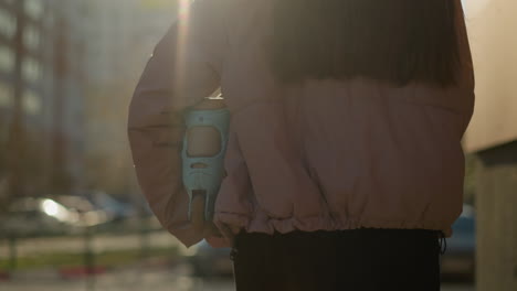 close-up of a girl with long brown hair flowing, wearing a peach jacket and holding a skateboard as she walks through an urban area at sunset, with warm light casting a serene glow on her surroundings