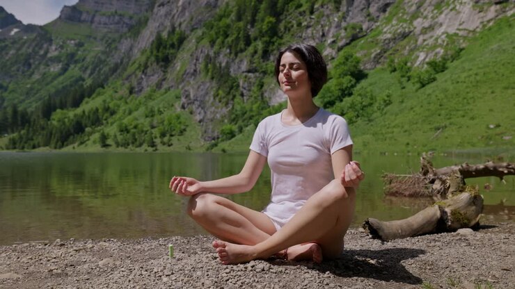Close up portrait of woman with short black hairs sitting on rocky shore of swiss lake and doing yoga exercise. Closed eyes.Dolly zoom out. Summer sunny day in Switzerland. European female model girl.
