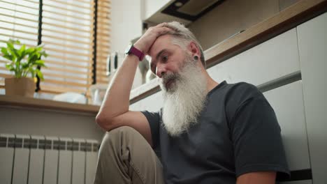 A-serious-man-with-gray-hair-and-a-lush-gray-beard-in-a-gray-T-shirt-sits-on-the-floor-and-holds-his-head-with-one-leg-in-the-kitchen.-A-thoughtful-and-serious-elderly-man-sits-in-the-kitchen-and-looks-at-the-floor-while-holding-his-forehead-with-his-hand