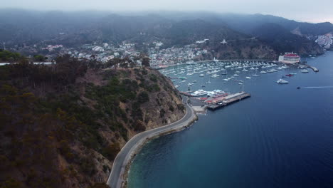 Schließende-Luftaufnahme-Der-Küstenstraße-Nach-Avalon-Bay-Hafen,-Casino-Und-Nebligen-Hügeln,-Kalifornien