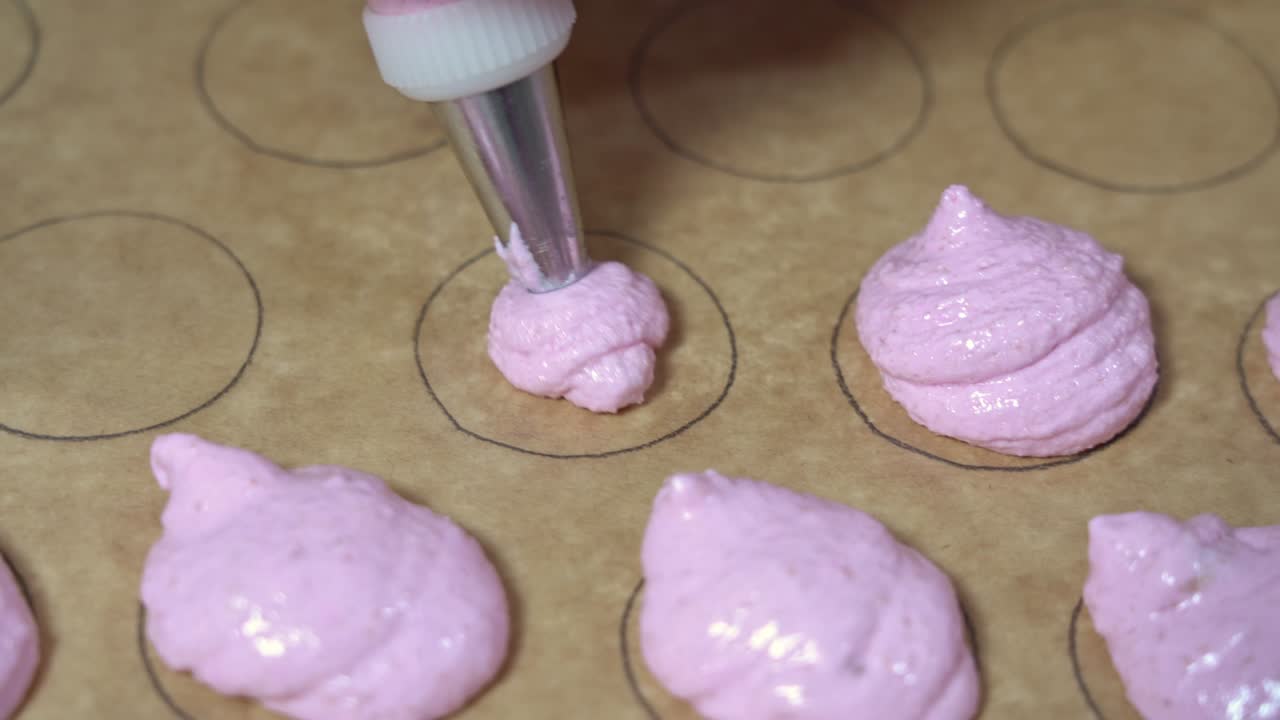 una toma de cerca de un panadero usando una bolsa de pastelería con masa rosa de macarrón para llenar formas circulares en papel de pergamino.