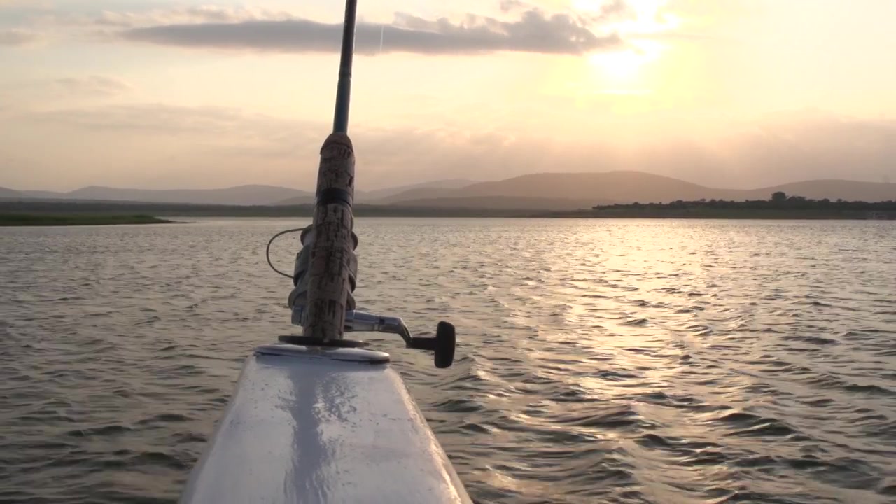 Bellissimo clip in prospettiva in prima persona di una canna da pesca montata sul ponte di una barca che naviga su acque agitate del lago verso un tramonto dorato e colline ondulate in lontananza nel Sudafrica rurale