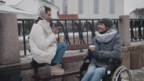 muslim woman and her disabled friend in wheelchair drinking takeaway coffe on a bench in city in winter 1