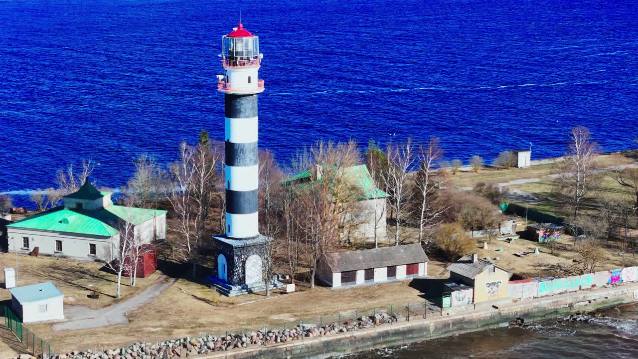 Aerial view of a striking black-and-white lighthouse standing by vivid blue river waters, surrounded by rustic buildings and bare trees along the rocky shoreline.