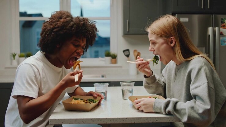 Lifting chopsticks, two women in casual clothing sharing to-go bowls in kitchen, exchanging looks