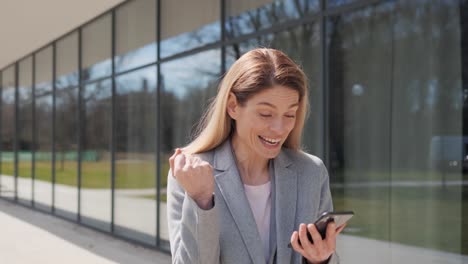 una mujer adulta emocionada se regocija con el mensaje en su teléfono inteligente. una mujer de negocios feliz mirando su teléfono celular celebra las buenas noticias. el éxito de la victoria. el retrato de un jugador afortunado mostrando el gesto del ganador del sí afuera.