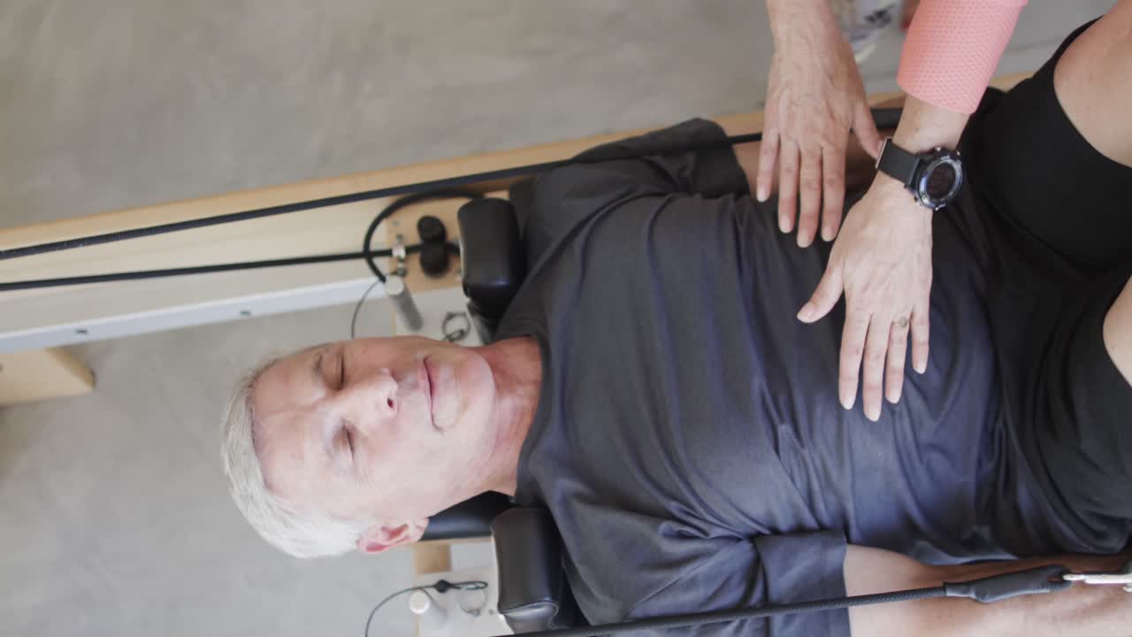 hombre caucásico anciano enfocado usando reformador con entrenadora de pilates femenina, sin alteración, en cámara lenta.