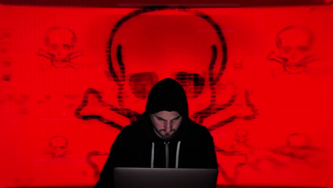 black hooded hacker programs computer virus in front of huge laughing green skull and crossbones symbol