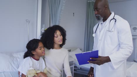 Médico-Senior-Afroamericano-Con-Portapapeles-Hablando-Con-Madre-E-Hija-En-El-Hospital