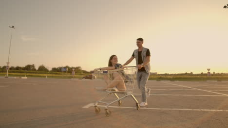 Cupé-Joven-Y-Elegante-Divirtiéndose-Montando-Con-Un-Carrito-De-Compras-En-El-Estacionamiento-Al-Aire-Libre-Cerca-Del-Supermercado