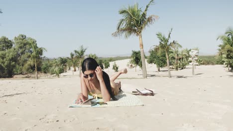 Fröhliche-Hispanische-Frau-Mit-Sonnenbrille,-Die-Am-Strand-Liegt-Und-Ein-Buch-In-Der-Sonne-Liest,-Kopierraum,-Zeitlupe