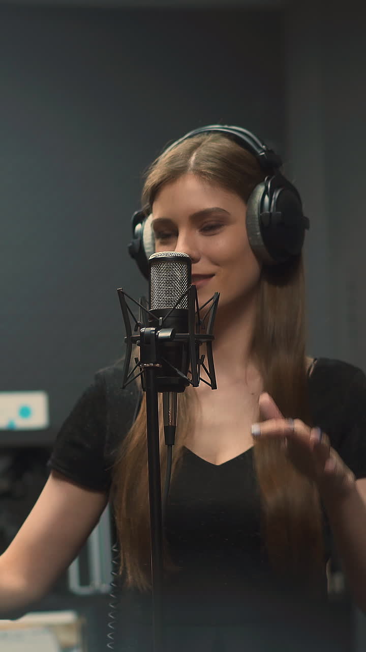 Attractive young blonde vocalist with headphones sings new song with modern microphone dancing against guitars hanging on wall in record studio closeup