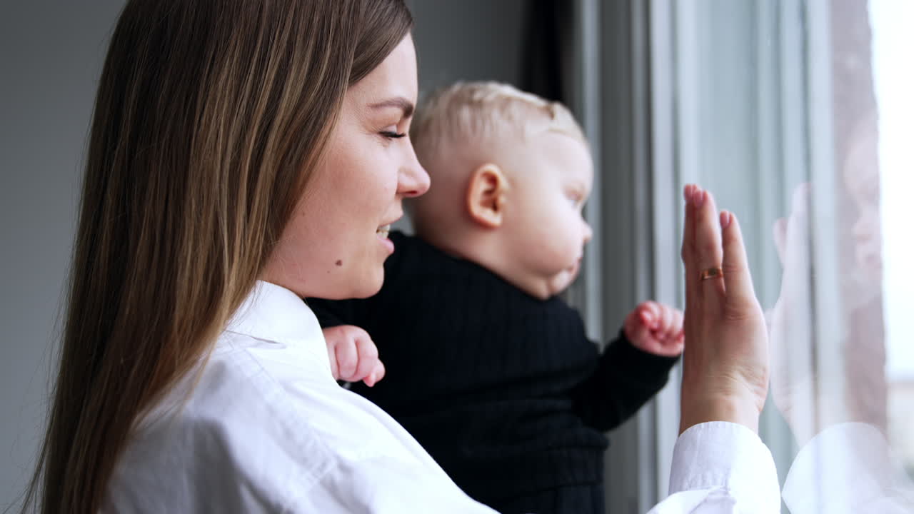 Long-haired Caucasian woman holding a chubby little baby. Mother and son looking in the window. Close up.