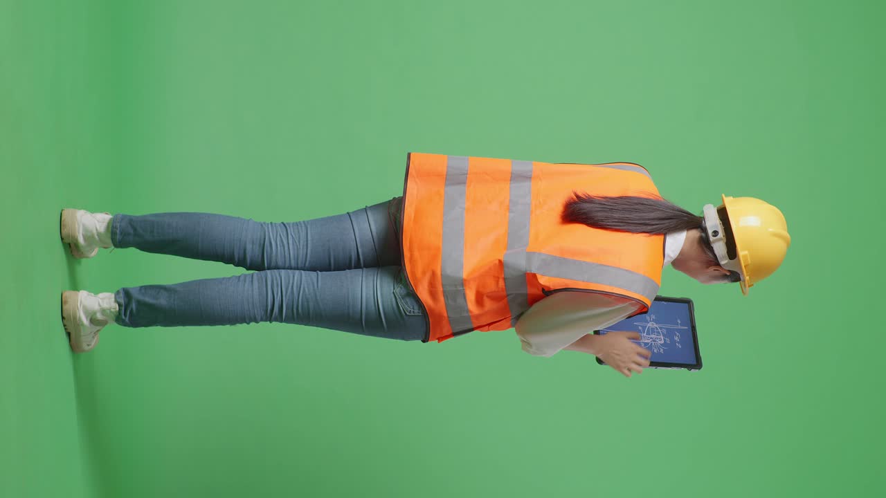 Full Body Back View Of Asian Female Engineer With Safety Helmet Looking At The Wind Turbine Blueprint On A Tablet While Standing In The Green Screen Background Studio