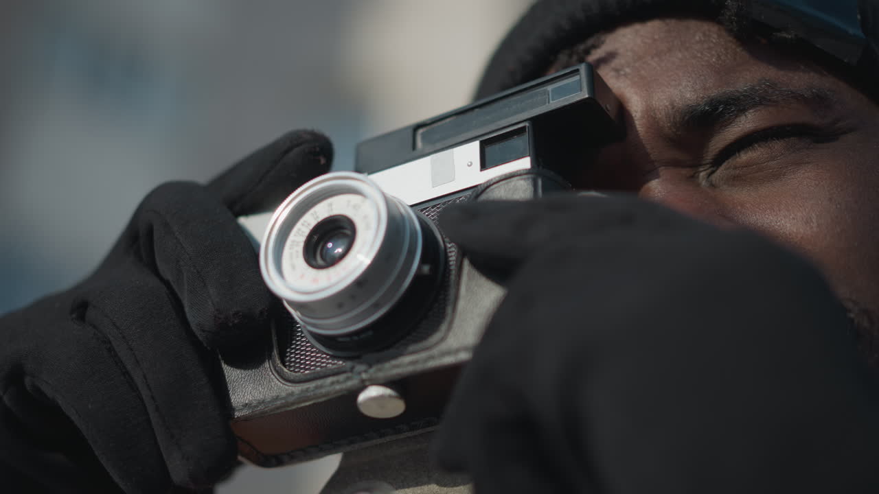 close up of cinematographer hands adjusting lens and pressing shutter trigger on digital camera, gloves protecting from cold, bright winter daylight illuminating snow covered street scene