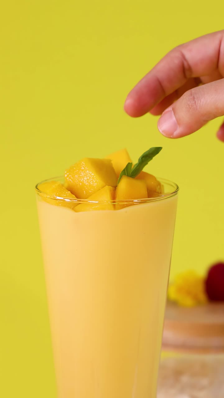 Bicchiere alto di smoothie al mango guarnito con foglie di menta, circondato da frutta fresca su sfondo luminoso.